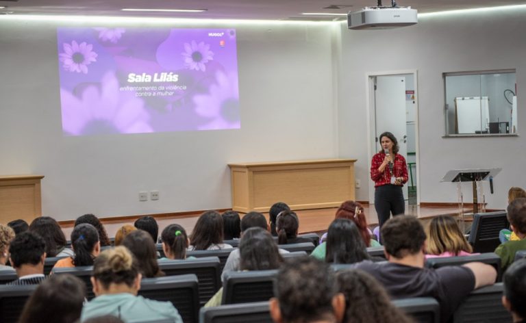O Hospital Estadual de Urgências Governador Otávio Lage de Siqueira (Hugol) inaugurou, nesta quinta-feira (28/05), sua Sala Lilás, espaço destinado ao acolhimento e apoio de mulheres vítimas de violência. A iniciativa tem como objetivo fortalecer a assistência humanizada e garantir um atendimento mais seguro, reservado e acolhedor às vítimas atendidas. A Sala Lilás foi estruturada para oferecer privacidade, escuta qualificada e suporte durante o atendimento multiprofissional. O ambiente também poderá ser utilizado em atendimentos que que também envolvam crianças e adolescentes em situação de vulnerabilidade. O evento reuniu 168 profissionais do hospital, entre equipes assistenciais, multiprofissionais e setores estratégicos, reforçando a importância da qualificação contínua para o atendimento humanizado e identificação precoce de situações de violência. A inauguração contou com palestra voltada aos profissionais, ministrada pela coordenadora-geral dos Ciclos de Vida e Violências na Superintendência de Políticas e Atenção Integral à Saúde da Secretaria da Saúde de Goiás, Lígia da Fonseca Bernardes. Ela abordou temas relacionados ao enfrentamento da violência contra a mulher, incluindo: fluxo de atendimento dos casos; violência de gênero; Lei Maria da Penha; tipos de violência; ciclo da violência; identificação de sinais de abuso e vulnerabilidade. Capacitção das equipes Segundo Lígia, a capacitação das equipes de saúde é fundamental para garantir acolhimento adequado às vítimas. “Os profissionais da saúde têm papel essencial na identificação precoce dos sinais de violência e no acolhimento humanizado dessas mulheres”. “Muitas vezes, o hospital é a primeira porta de apoio procurada pela vítima, e estar preparado para esse atendimento pode fazer toda a diferença no rompimento do ciclo da violência”, destacou. Sala Lilás – avanço no cuidado De acordo com a supervisora multiprofissional do Hugol, Gabriela Vieira, a implantação da Sala Lilás representa um avanço importante no cuidado às vítimas em situação de vulnerabilidade. “A criação desse espaço fortalece o acolhimento humanizado dentro da unidade e proporciona mais segurança, privacidade e suporte emocional às pacientes atendidas. É um ambiente pensado para oferecer escuta qualificada e assistência integral”, afirmou. A criação da Sala Lilás integra as ações de fortalecimento das políticas de proteção e cuidado às vítimas de violência desenvolvidas pelo Hugol, promovendo um ambiente mais acolhedor e preparado para assistência integral aos pacientes em situação de vulnerabilidade. Além do acolhimento imediato, o espaço também busca contribuir para o encaminhamento adequado das vítimas à rede de apoio e proteção, fortalecendo o trabalho integrado entre saúde, assistência social e segurança pública. Saiba mais Sala Lilás do Hugol vai acolher mulheres vítimas de violência